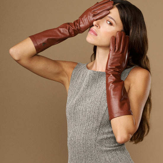 Cognac leather below-elbow gloves with curved fit, hands posed near head